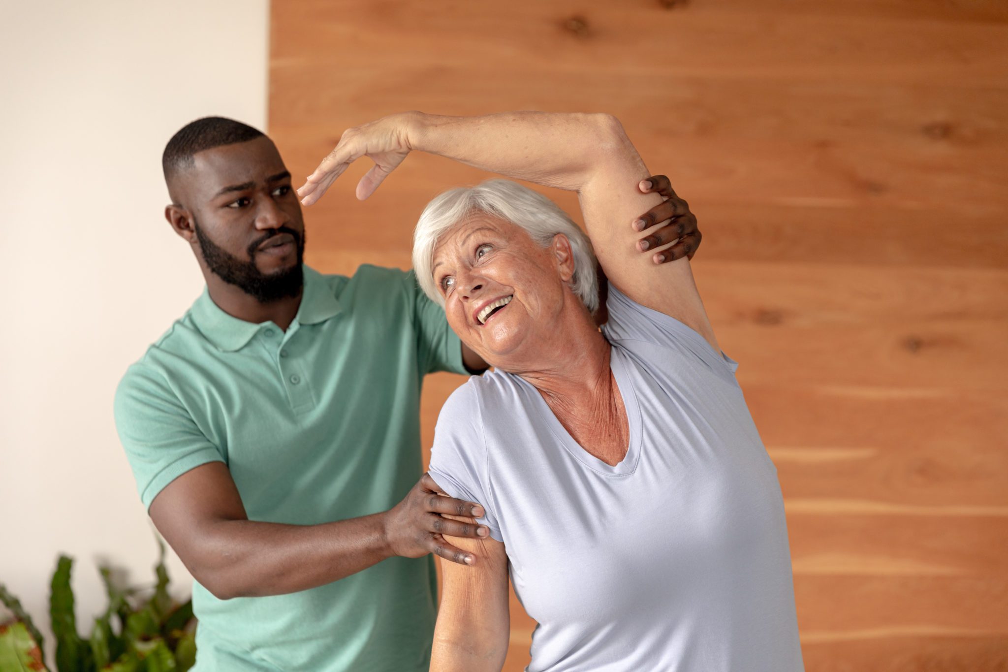 African american male physiotherapist helping caucasian senior woman to stretch at home. Physiotherapy and rehabilitation concept